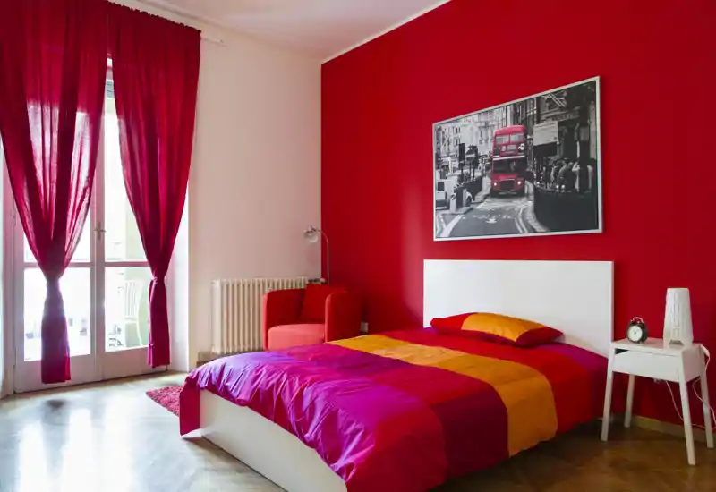 Bright, modern bedroom with a double bed dressed in vivid red and orange linens, a bedside table and an armchair beside a tall window leading to a balcony.