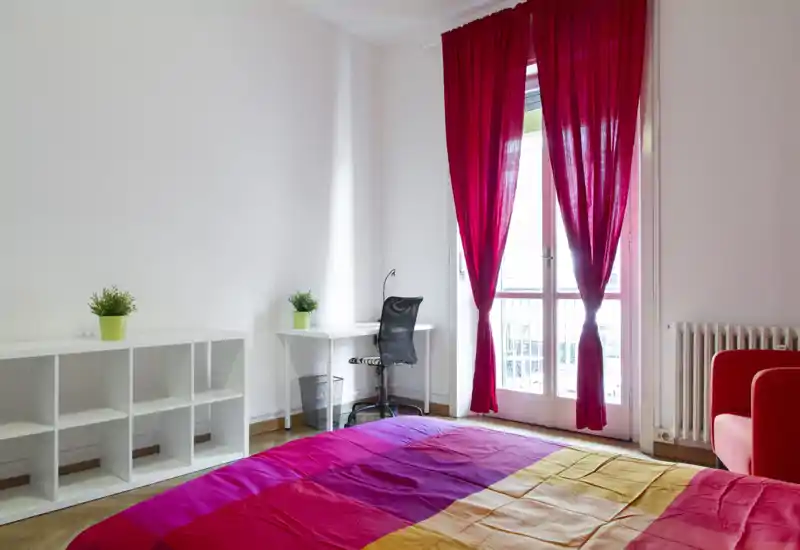 Bright, colorful bedroom with a large bed in the foreground, a desk and chair by the balcony door, and vibrant magenta curtains letting in natural light.