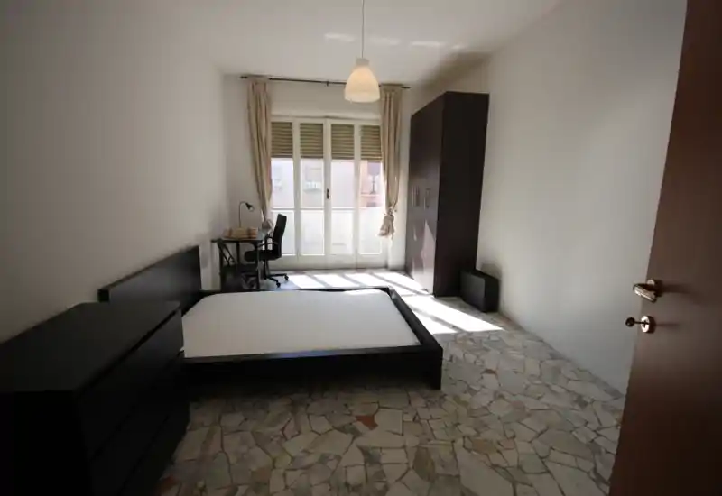 Camera da letto luminosa e arredata in modo essenziale con struttura letto matrimoniale, comò, armadio e una scrivania vicino alla grande porta finestra che lascia entrare molta luce naturale.