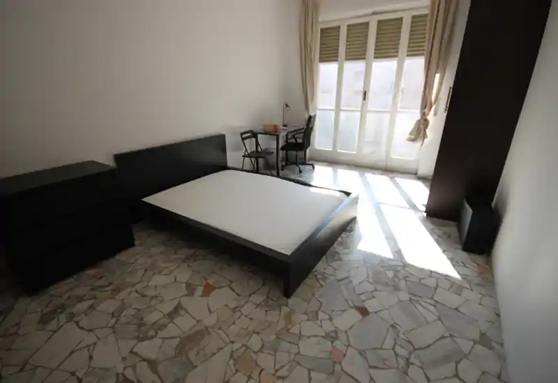 Camera da letto luminosa e minimal con letto basso in legno nero, comò coordinato e una piccola scrivania vicino a grandi finestre che lasciano entrare molta luce naturale.