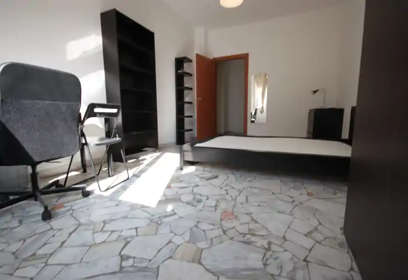 Camera da letto luminosa e arredata in modo semplice con struttura letto bassa nera, materasso e scaffalature pratiche. La luce naturale inonda la stanza e sono presenti una sedia da studio e un mobile per riporre.