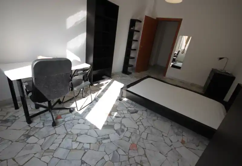 Camera da letto minimalista e luminosa con struttura del letto bassa nera, scrivania e scaffali; la luce naturale mette in risalto il pavimento in mosaico e gli arredi moderni e semplici.