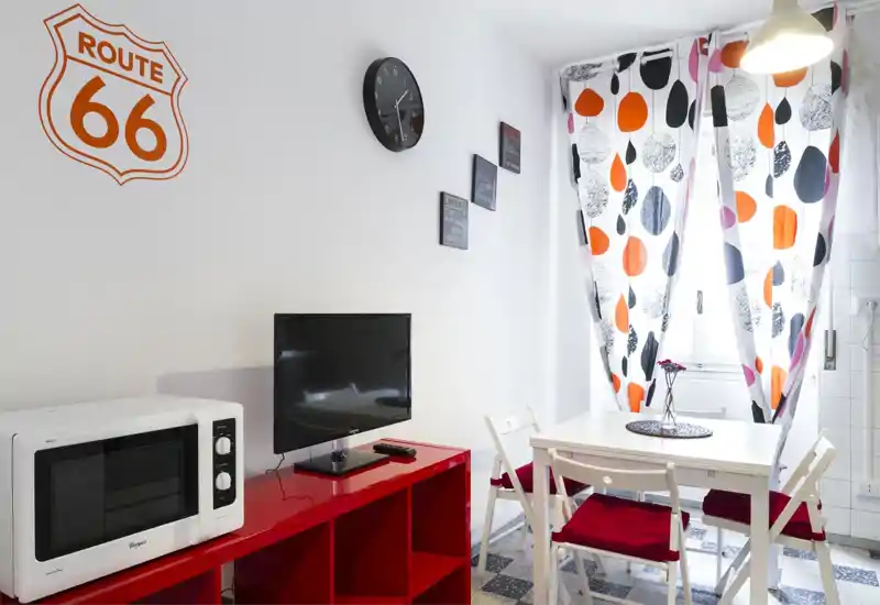 Soggiorno/pranzo compatto con tavolo bianco, sedie pieghevoli con cuscini rossi, tende a fantasia e angolo multimediale con TV e microonde — luminoso, moderno e arredato con efficienza.