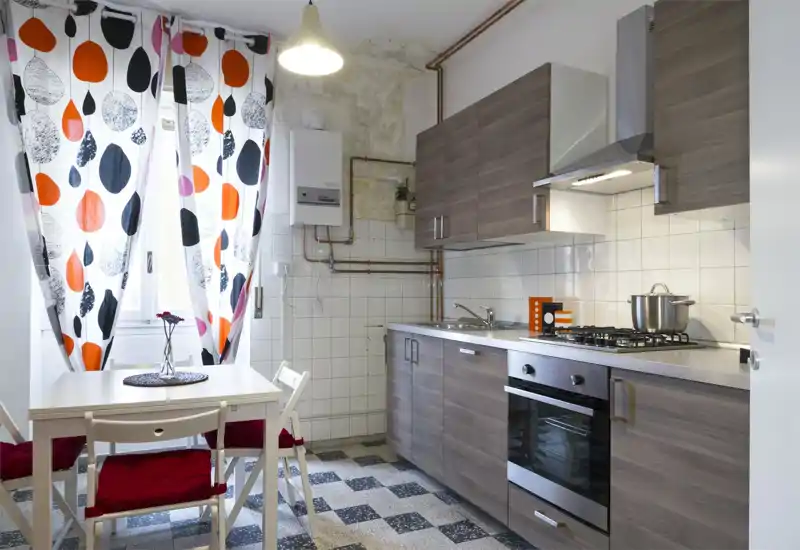 Cucina compatta e luminosa con mobili effetto legno, piano cottura a gas, forno incassato e un piccolo tavolo da pranzo vicino alla finestra con tende colorate.