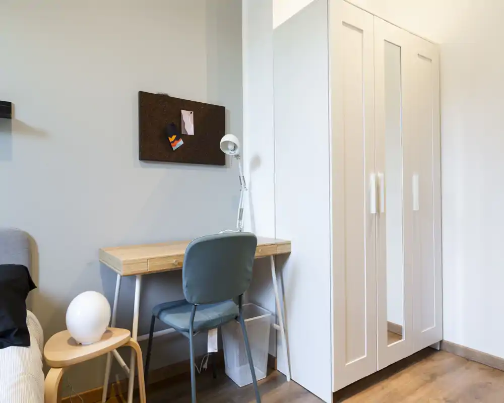 Rincón de dormitorio luminoso y minimalista con un pequeño escritorio de madera, silla tapizada y un armario blanco. Se aprecia el borde de la cama y una zona de trabajo compacta ideal para una persona.