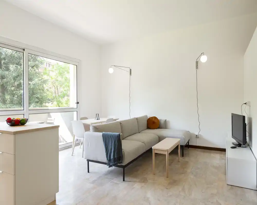Salón moderno y luminoso con sofá en L gris claro, mesita de centro y TV; gran ventana con vistas al verde que llena el espacio de luz natural.
