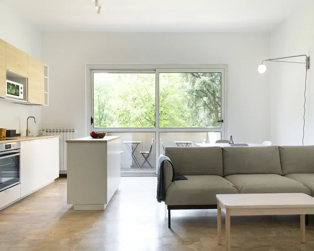 Espacio de sala moderno y luminoso de planta abierta con sofá confortable, mesa de centro y una isla de cocina adyacente que se abre a un balcón con vegetación.