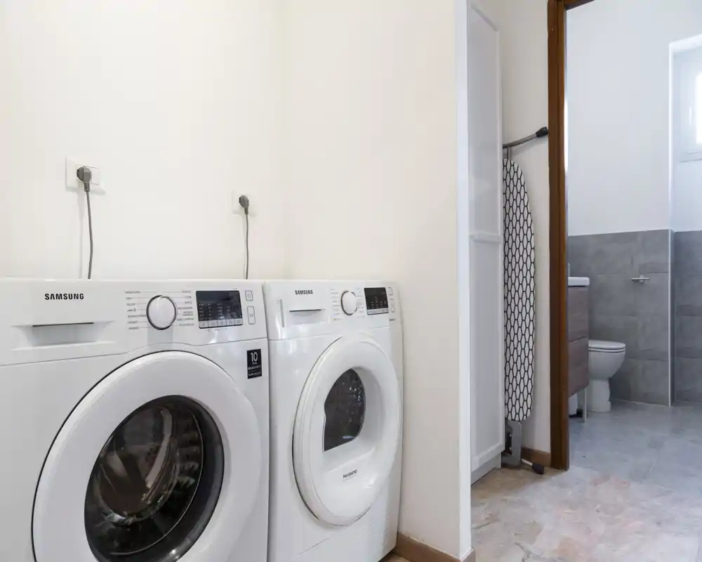 Rincón de lavandería luminoso con lavadora y secadora colocadas lado a lado, junto a una puerta que conduce a un baño con azulejos.
