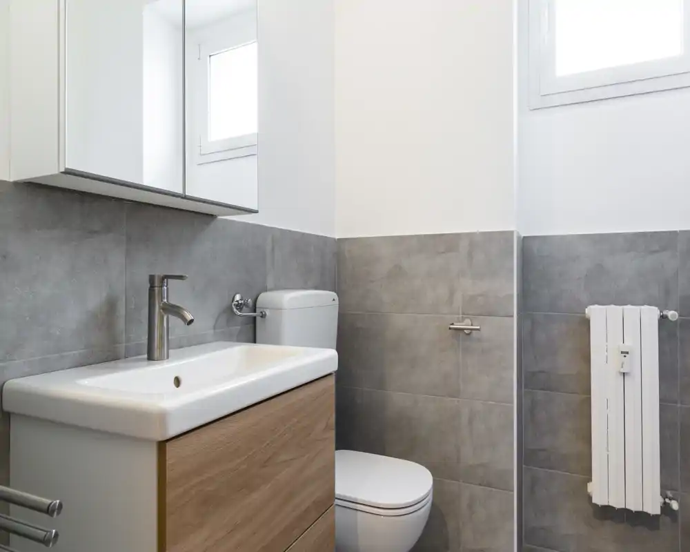 Baño moderno y compacto con armario con espejo, lavabo rectangular sobre mueble efecto madera y inodoro; azulejos grises y una pequeña ventana que aporta luz natural.