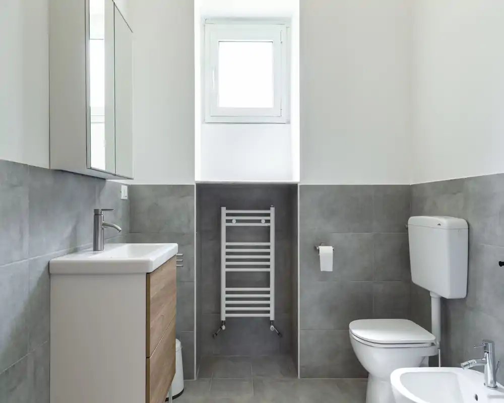 Baño moderno y limpio con toallero calefactado de pared, lavabo con mueble, inodoro y bidé en un espacio luminoso de tonos neutros.