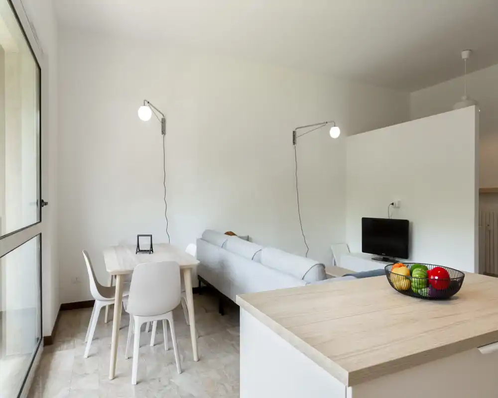 Espacio de estar moderno y luminoso con sofá gris claro, pequeña mesa de comedor y gran ventana que aporta luz natural — ideal para un apartamento urbano con estilo.