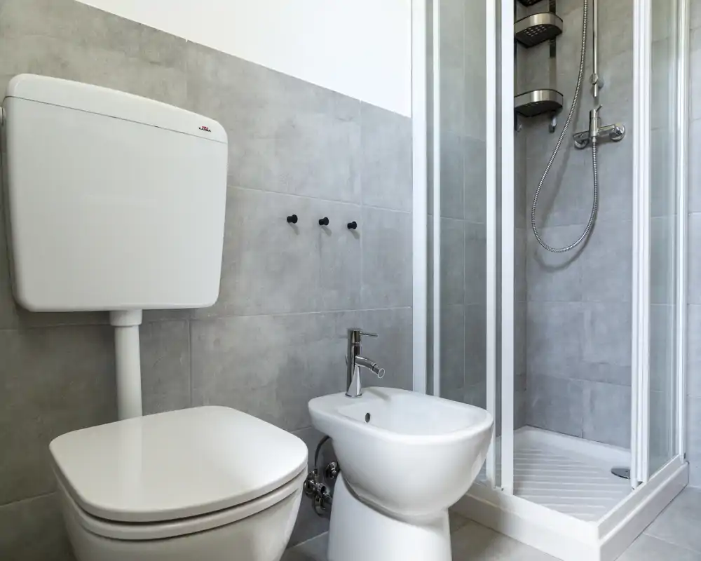 Baño moderno y compacto con inodoro, bidé y ducha en esquina; azulejos grises neutras y grifería limpia que crean un espacio funcional y fresco.