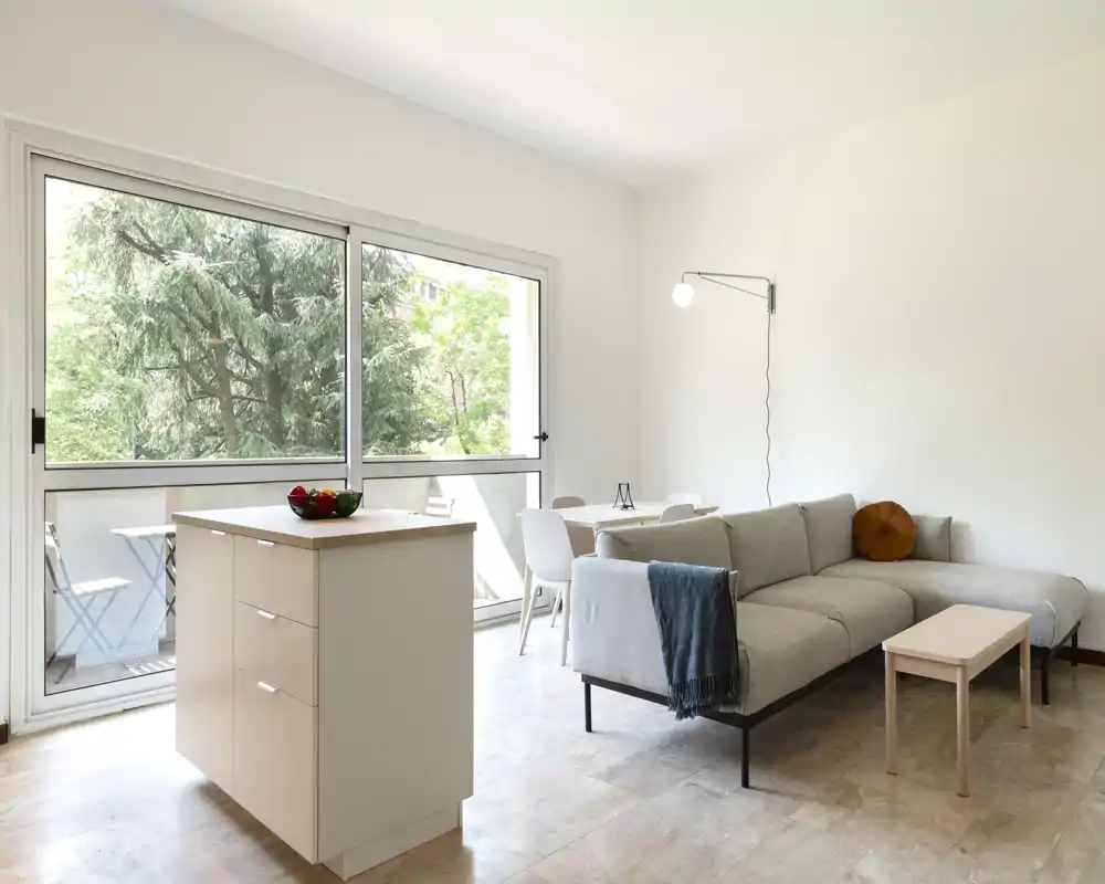 Salón moderno y luminoso con sofá esquinero gris claro, mesa de centro pequeña y comedor junto a grandes ventanales con vistas a la vegetación — aireado y minimalista.