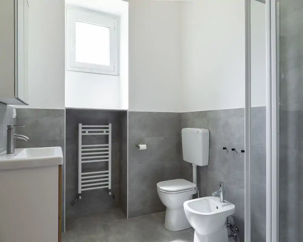 Baño moderno y luminoso con radiador mural, inodoro, bidé y lavabo; azulejos grises neutras y una ventana que aporta luz natural.