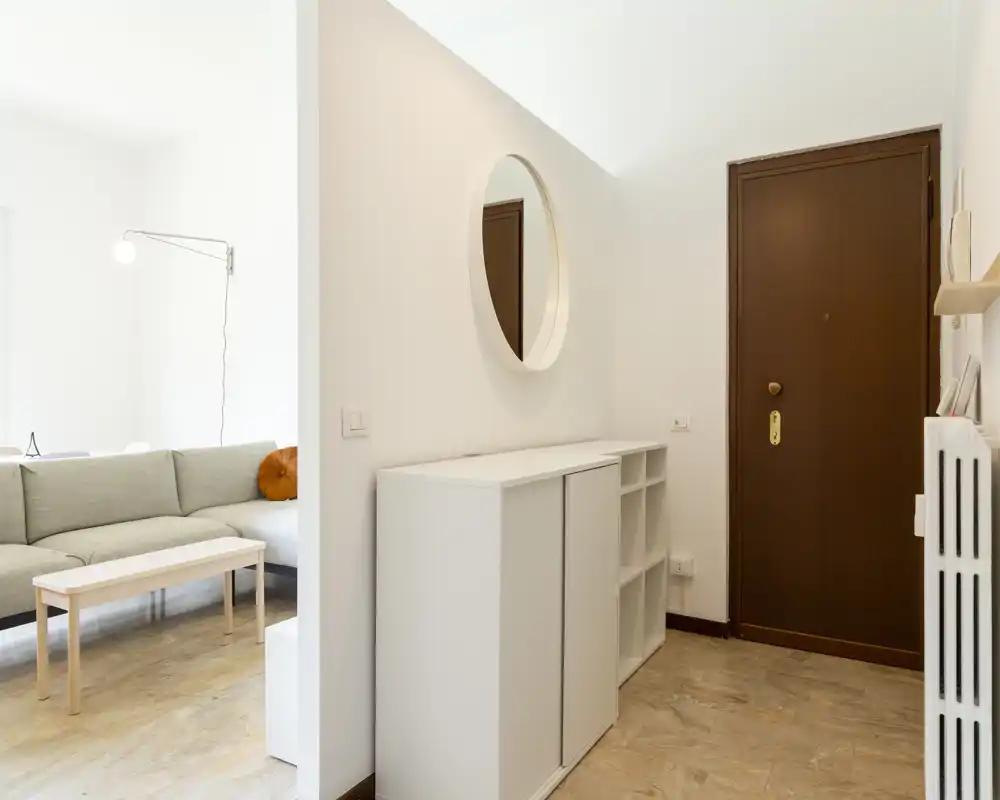 Entrada luminosa y minimalista con consola blanca, espejo redondo y puerta principal de madera; se ve la sala con sofá detrás del tabique.