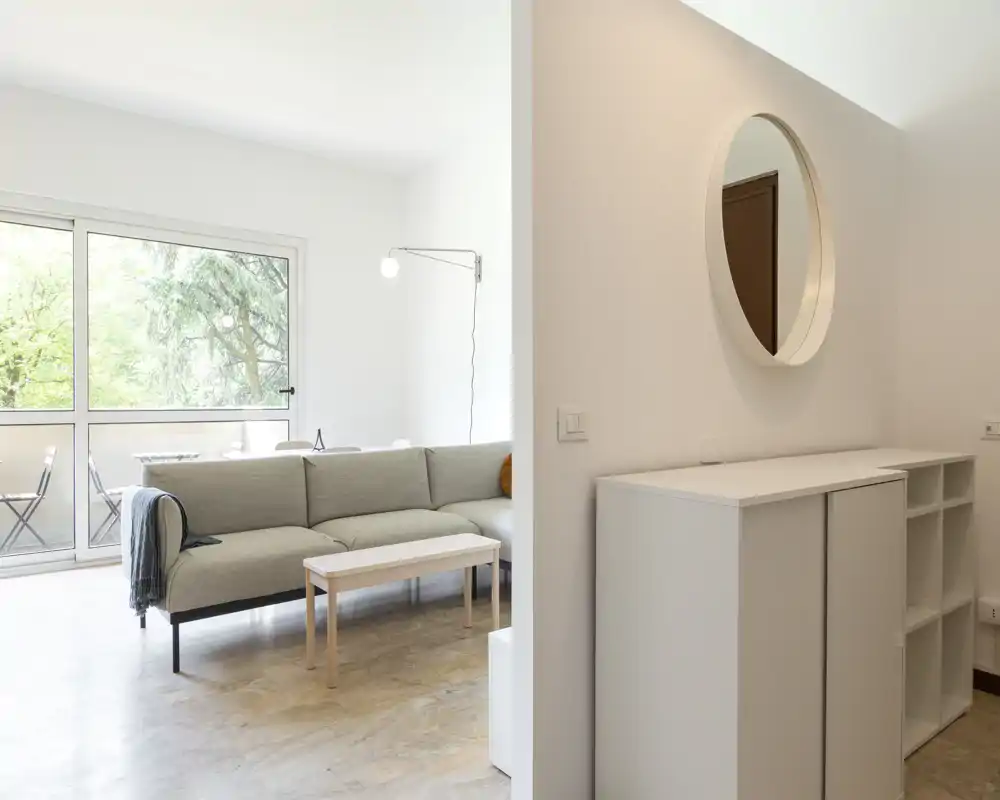 Sala de estar moderna y luminosa con sofá gris claro, mesa de centro y una gran ventana que da al balcón, ambiente relajado y ventilado.