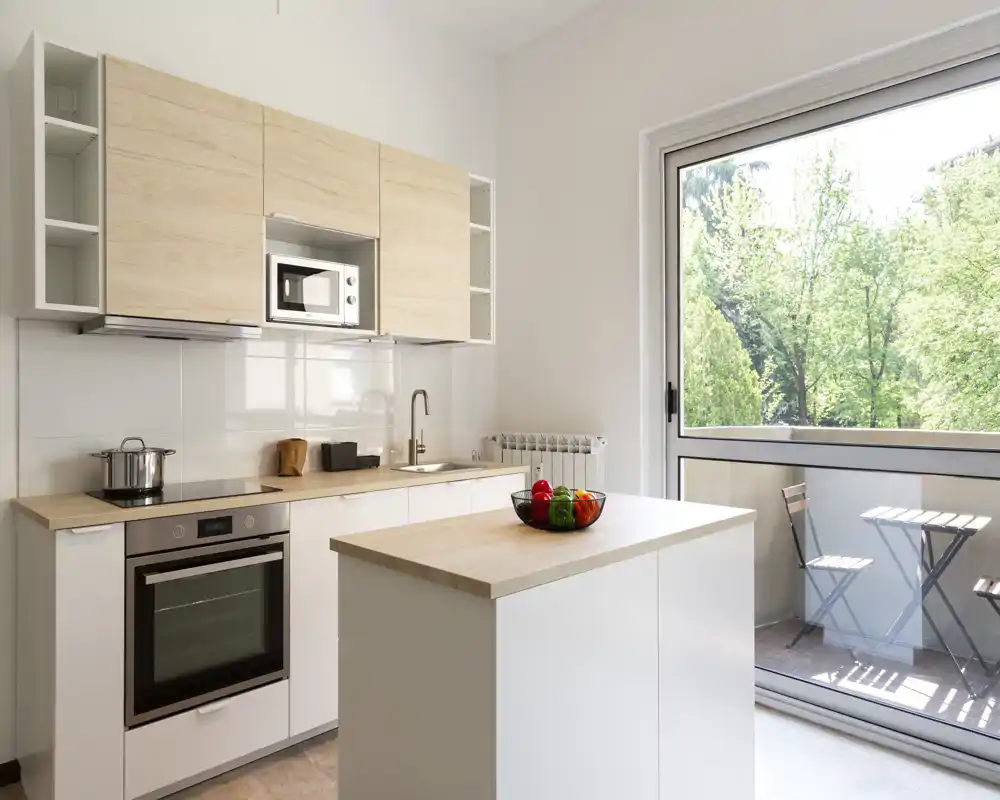 Cocina moderna y luminosa con isla central, horno empotrado y frentes de madera. Gran ventanal corredero se abre a un pequeño balcón que aporta mucha luz natural.