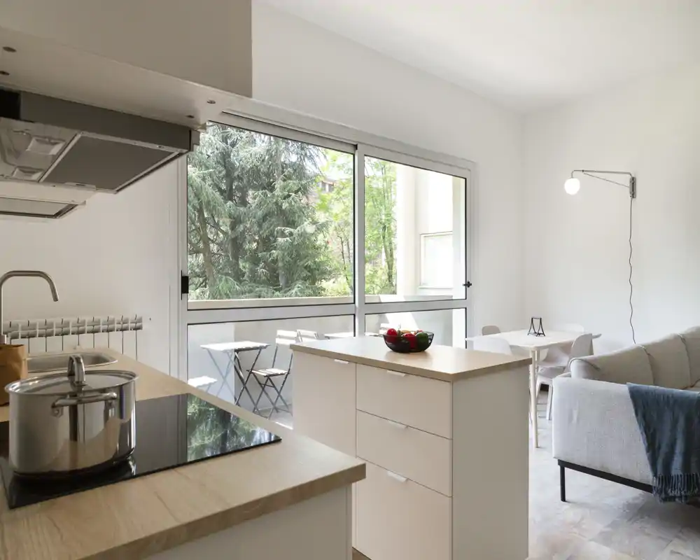 Zona de estar de planta abierta y luminosa con cocina moderna, sofá acogedor y una pequeña mesa de comedor junto a grandes ventanales con vistas al verde.