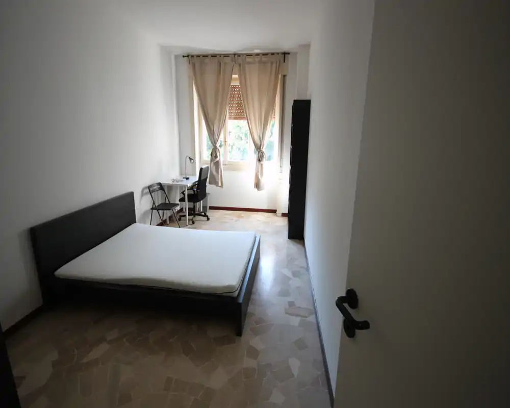 Camera da letto semplice e luminosa con letto matrimoniale, scrivania e luce naturale dalla finestra con tende leggere — spazio pulito e funzionale ideale per studenti o professionisti.