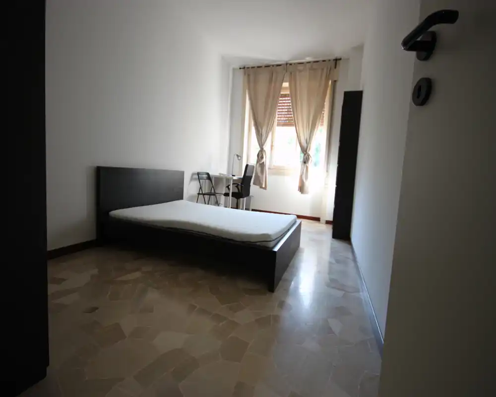 Camera da letto minimal e luminosa con letto basso matrimoniale, scrivania vicino alla finestra e arredo neutro. La luce naturale mette in risalto il pavimento a effetto pietra.