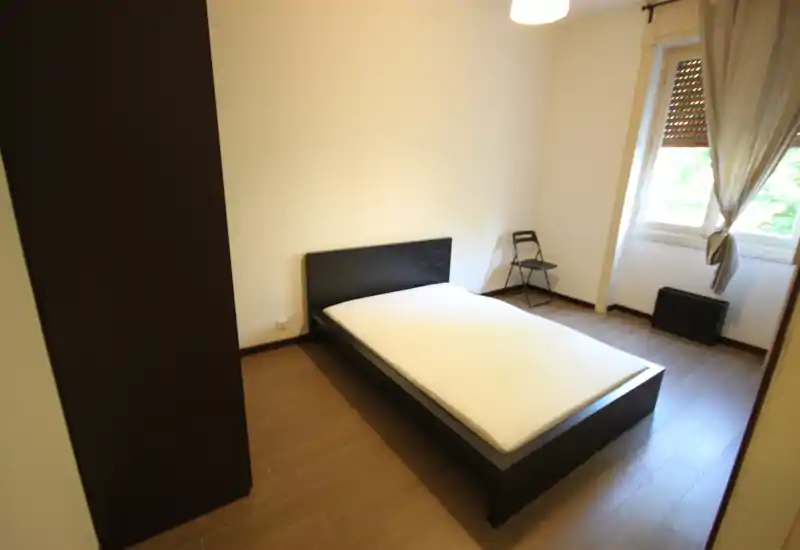 Camera da letto semplice e luminosa con letto matrimoniale su struttura in legno scuro, armadio a sinistra e finestra con tende che garantisce luce naturale. Minimalista e ordinata, adatta agli annunci di affitto.