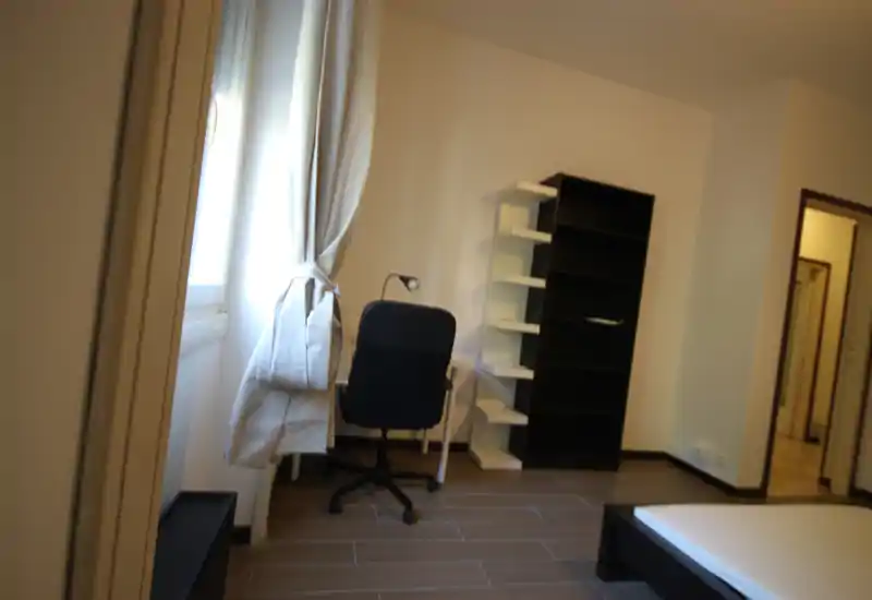 Camera da letto minimale con letto basso parzialmente visibile, scrivania e sedia vicino alla finestra, libreria e arredo neutro; luce naturale brillante dalla finestra.