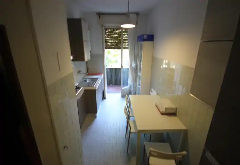 Cocina estrecha con zona de trabajo compacta, fregadero y cocina a la izquierda, mesa con sillas a la derecha y puerta al balcón que aporta luz natural.