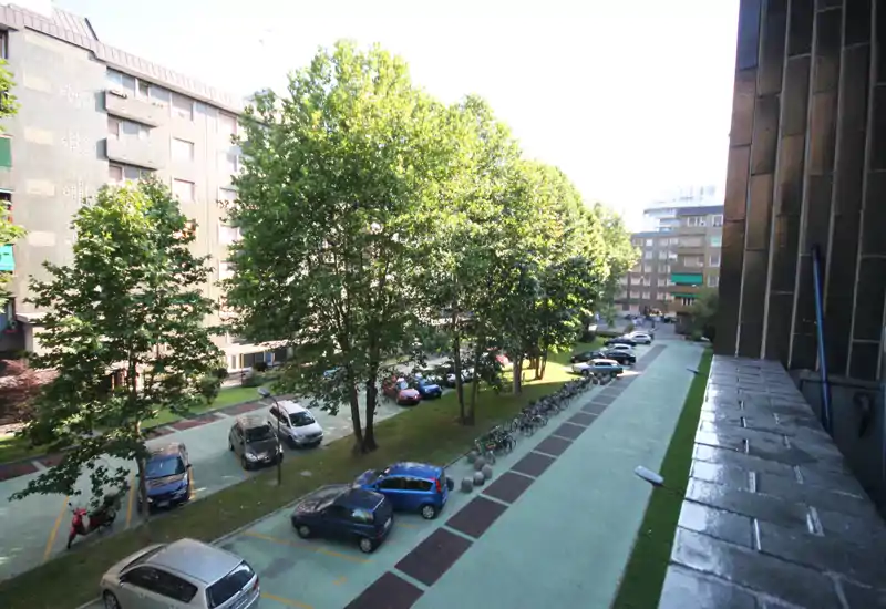 Cortile soleggiato con parcheggio alberato e rastrelliera per biciclette, che mostra gli spazi esterni disponibili e l'accesso dal palazzo.