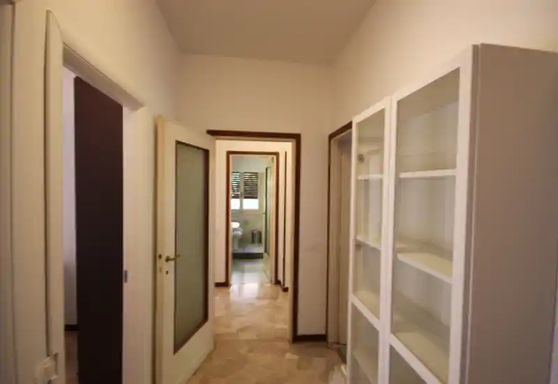 Corridoio/ingresso luminoso con pavimento in piastrelle chiaro, pareti bianche e mobile contenitore; la visuale attraversa più porte fino al bagno in fondo.