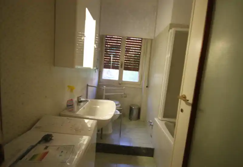 Bagno con lavabo, bidet, vasca con parete in vetro e una lavatrice; luce naturale dalla finestra che rende l'ambiente chiaro e funzionale.