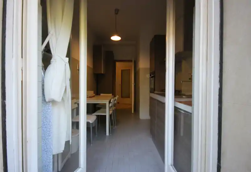 Cucina a corridoio luminosa con mobili in legno e un piccolo tavolo da pranzo; vista da una porta aperta che mostra una disposizione stretta e ordinata, ideale per appartamenti in città.