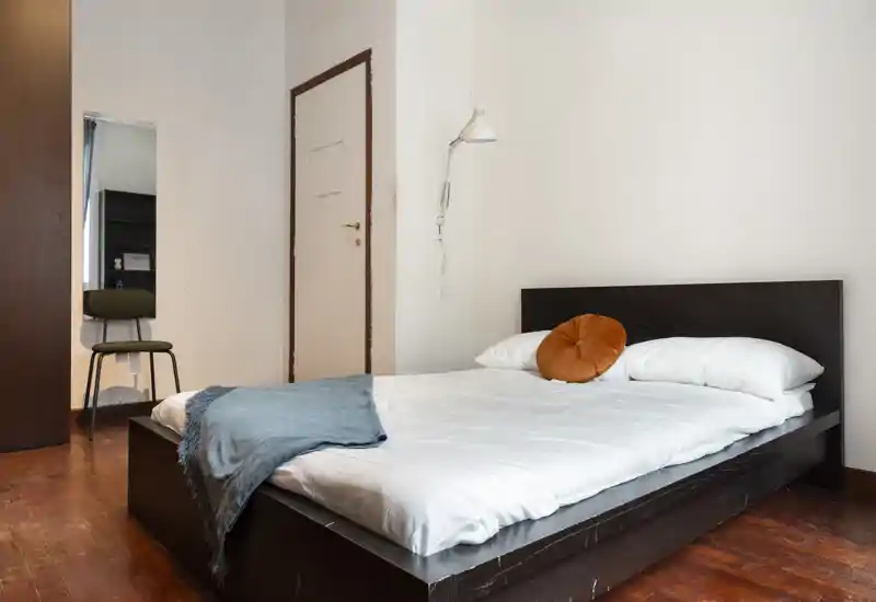 Dormitorio luminoso y sencillo con cama baja de madera oscura, ropa blanca, un cojín decorativo naranja y una manta azul. Suelo de madera, lámpara de pared y pequeño tocador visible al fondo.