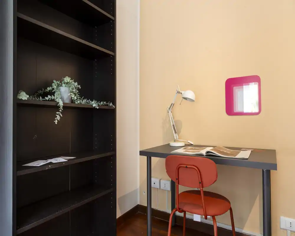 Rincón de estudio compacto con estantería oscura, mesa sencilla, silla roja y lámpara de escritorio — luminoso, ordenado y listo para trabajar.