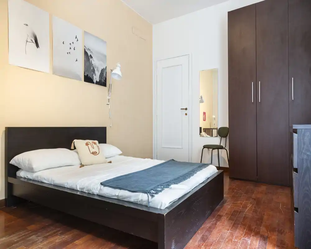 Dormitorio luminoso y moderno con cama baja de madera oscura, ropa de cama suave y manta decorativa. Suelo de madera cálido, armario alto y arte de pared sencillo que crean un ambiente acogedor y contemporáneo.