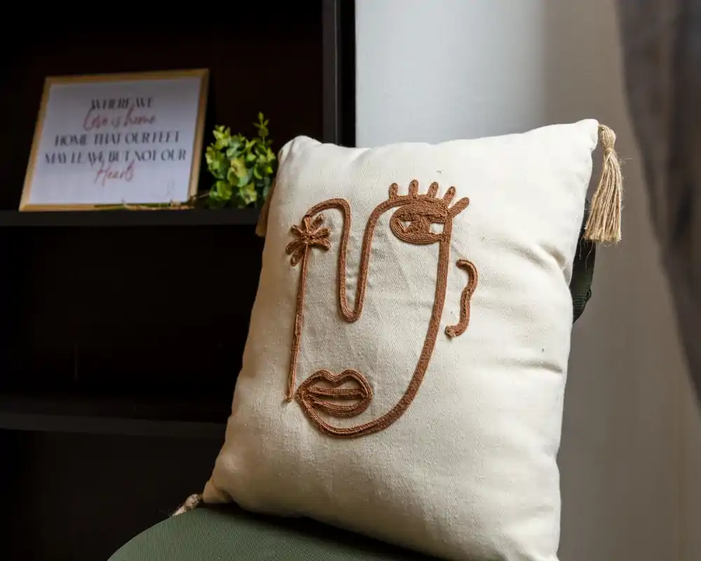 Primo piano di un cuscino decorativo con ricamo raffigurante un volto astratto appoggiato su una sedia verde, con una mensola scura e una stampa incorniciata sfocate sullo sfondo.