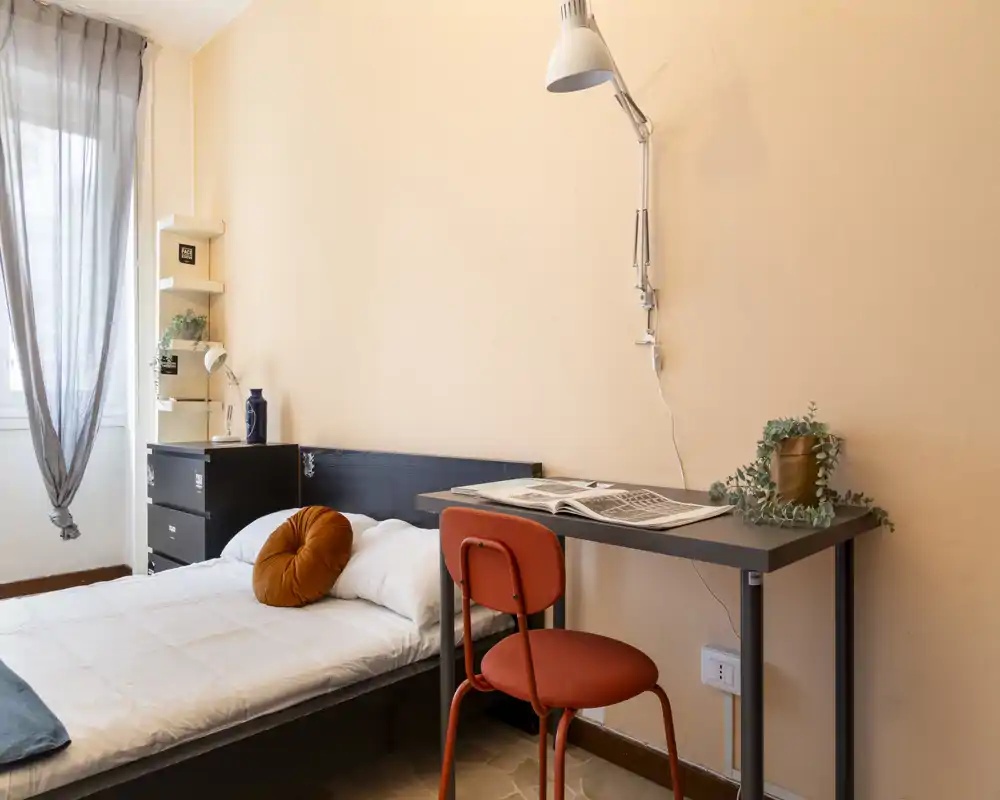 Camera da letto singola luminosa e accogliente con letto ben rifatto, scrivania compatta e sedia, e finestra che illumina la stanza — ideale per uno studente o una persona sola.