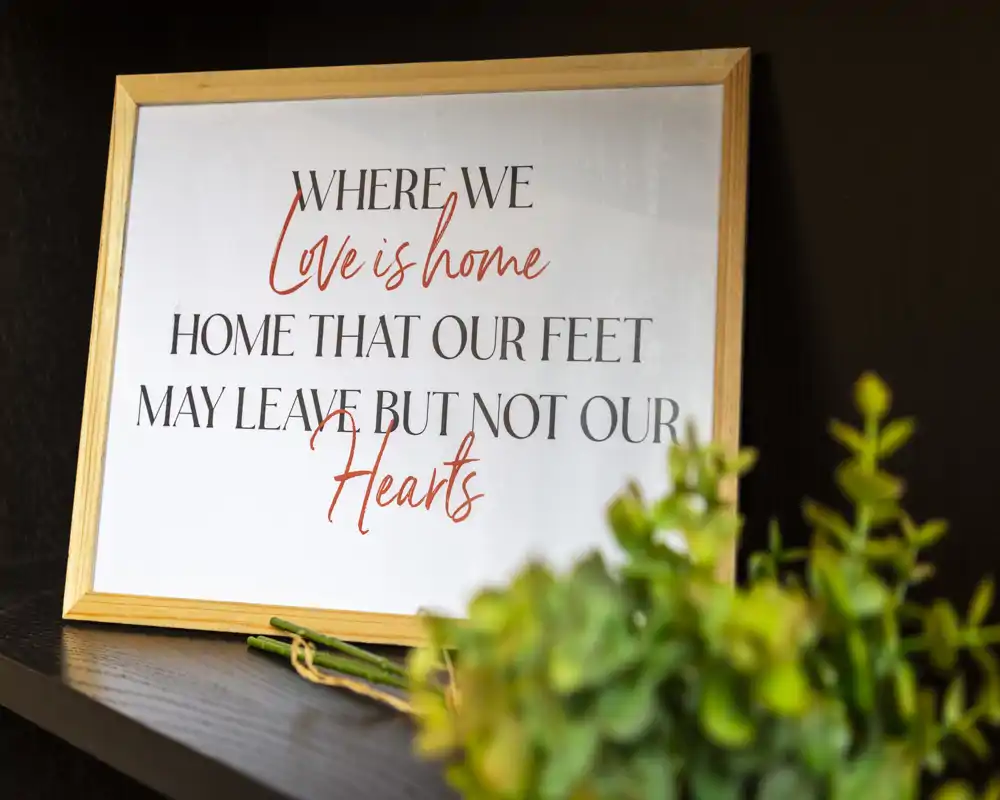 Primo piano di una cornice decorativa con la scritta "Where we live is home, home that our feet may leave but not our hearts" su una mensola scura; una piccola pianta verde è sfocata in primo piano.