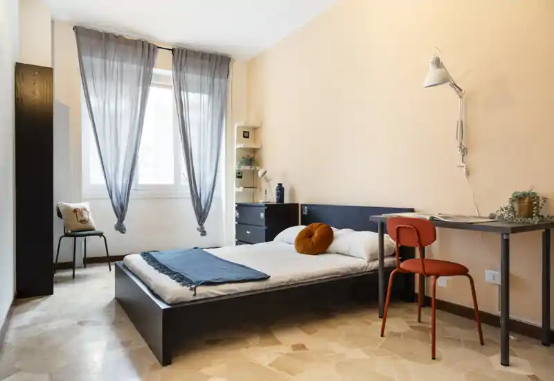 Camera da letto luminosa e ben arredata con letto basso matrimoniale, scrivania semplice e luce naturale morbida da una grande finestra—ideale per un soggiorno confortevole e minimalista.