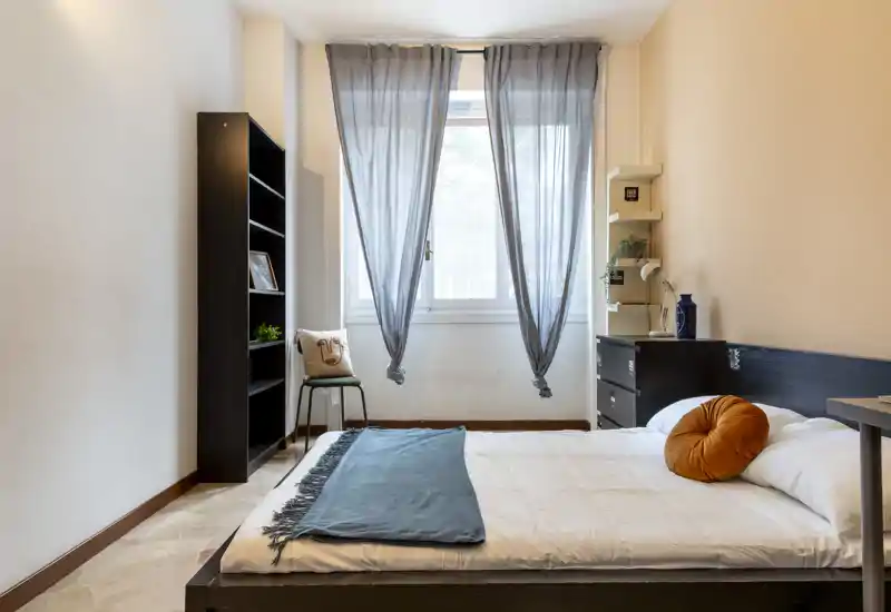 Camera da letto luminosa e ordinata con letto basso matrimoniale, cuscino decorativo e finestra con tende leggere che diffondono la luce naturale.