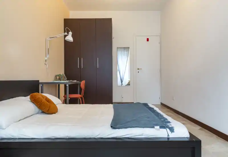 Camera da letto spaziosa e ben allestita con un grande letto in primo piano, un armadio in legno scuro, una scrivania e tessili caldi che creano un'atmosfera accogliente e minimale.