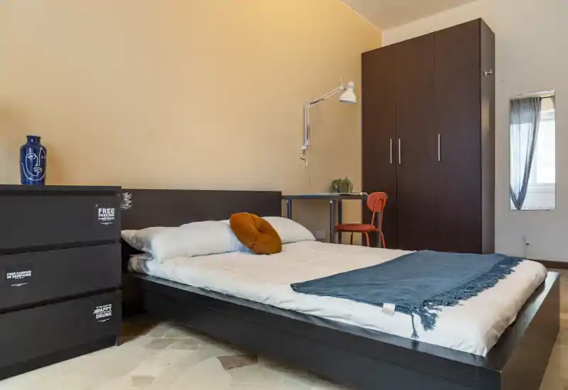 Camera da letto luminosa e ben arredata con letto basso in legno scuro, cuscini accoglienti e plaid; presente armadio e una scrivania, ideale per un soggiorno confortevole.