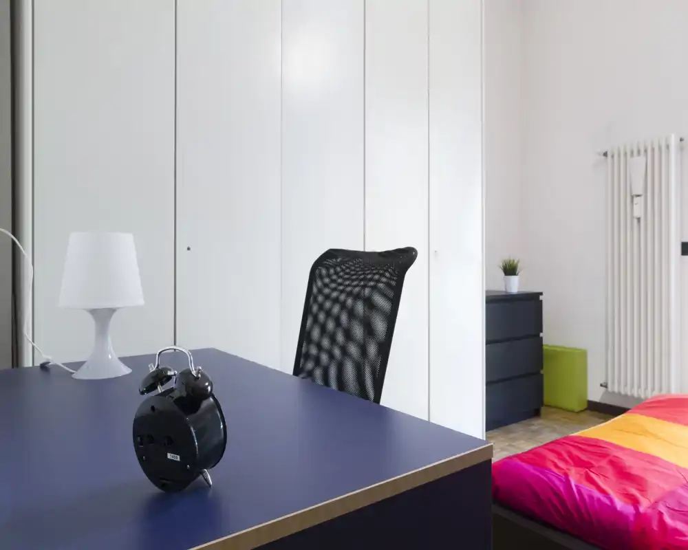 Rincón de dormitorio moderno y luminoso con un edredón colorido a la derecha, escritorio con lámpara y despertador en primer plano y armarios empotrados blancos al fondo.