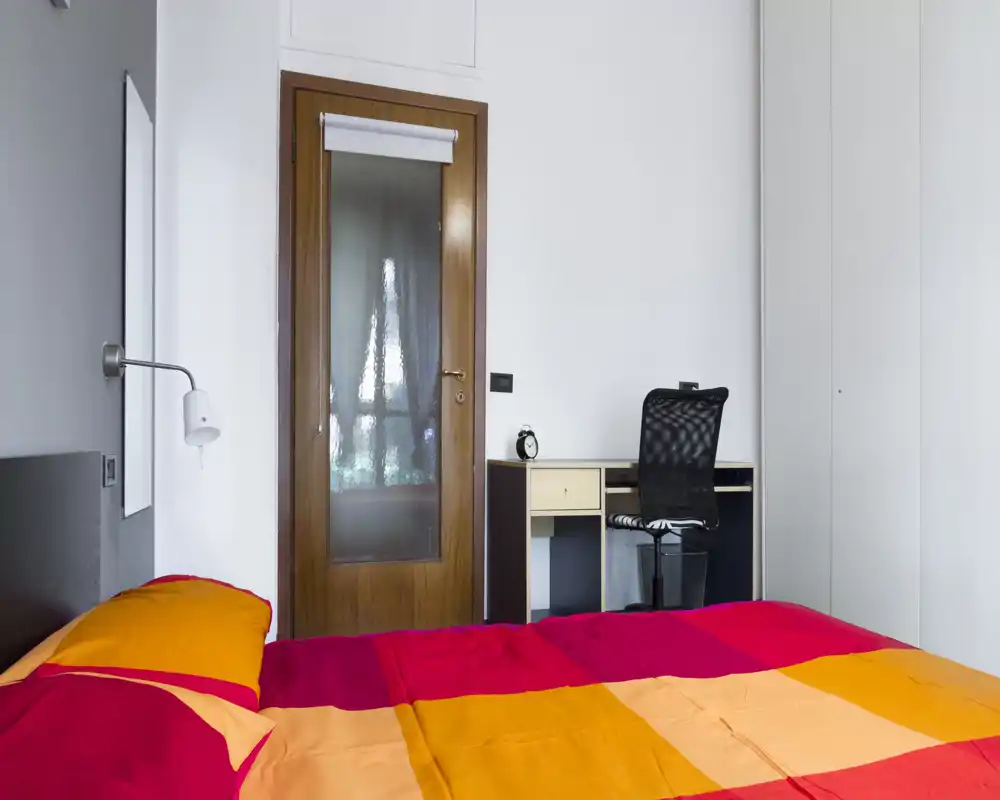Dormitorio luminoso con una cama doble de colores vivos en primer plano, un pequeño escritorio y silla en la pared y una puerta de madera con cristal esmerilado. Espacio sencillo y bien iluminado, apto para anuncios de alquiler.