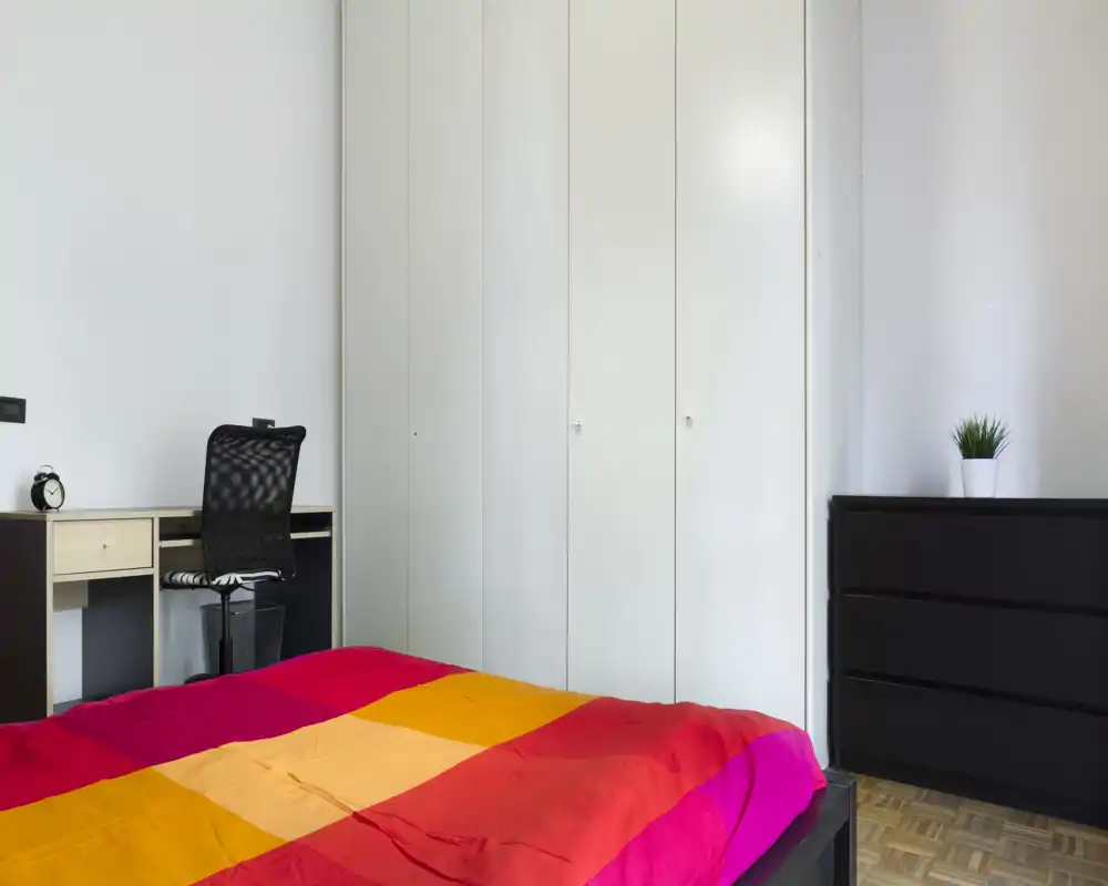Dormitorio moderno y luminoso con cama doble de colores, armario empotrado blanco, pequeño escritorio y cómoda; distribución sencilla y bien iluminada, ideal para anuncios.