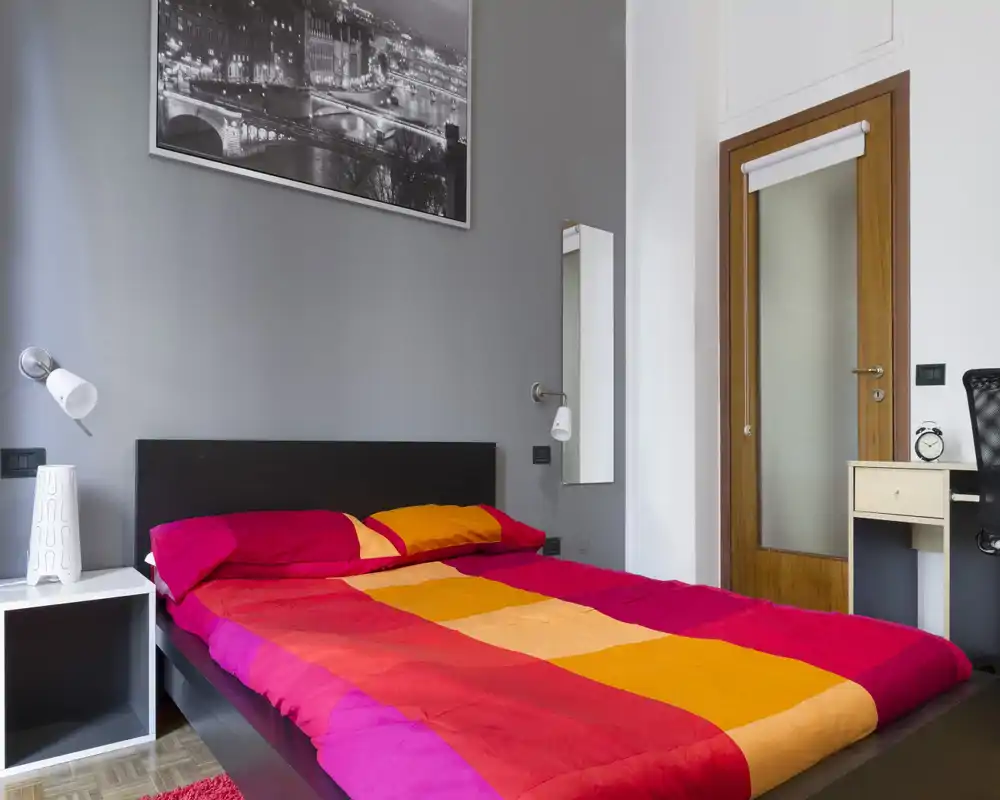 Dormitorio moderno y luminoso con cama doble con ropa de cama roja y naranja intensa, mesitas minimalistas, lámparas de pared y un pequeño escritorio junto a la puerta. Líneas limpias y colores llamativos crean un ambiente contemporáneo y acogedor, perfecto para anuncios.