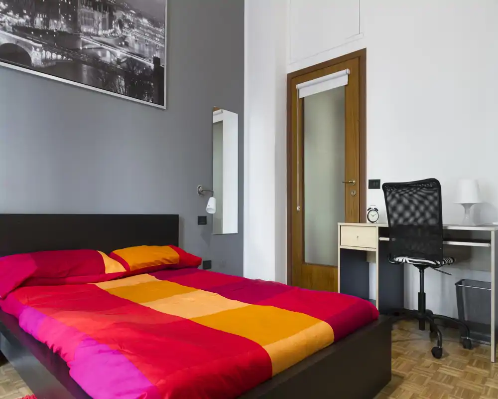 Dormitorio moderno y luminoso con una gran cama doble con ropa de cama colorida, un pequeño escritorio con silla junto a la puerta y un cuadro decorativo.