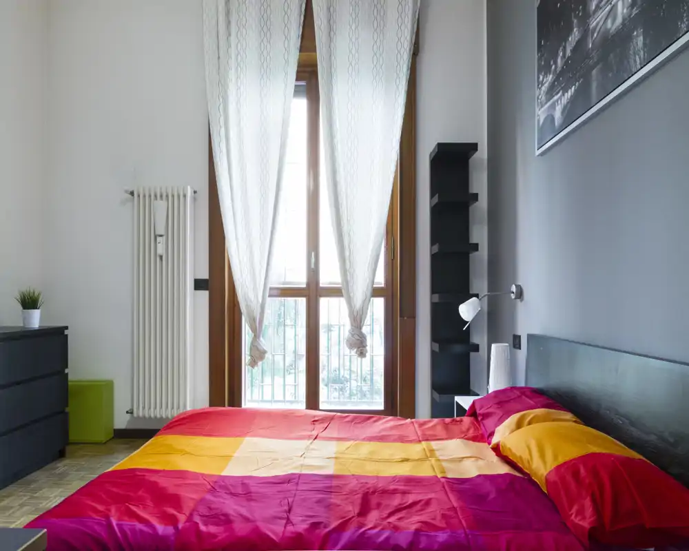 Dormitorio luminoso y bien decorado con una gran cama doble de ropa de cama colorida, ventana con cortinas traslúcidas que aporta luz natural y mobiliario minimalista.