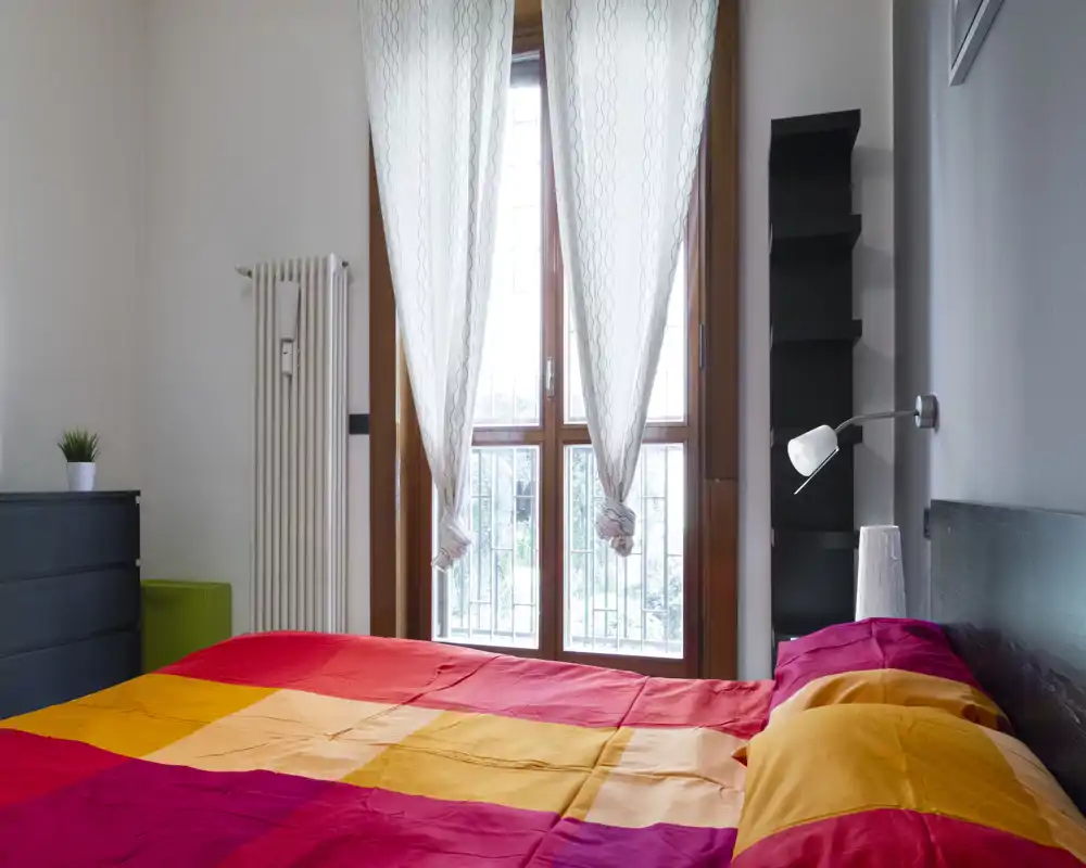 Dormitorio luminoso y ordenado con una cama grande y colorida frente a una ventana alta que deja entrar luz natural — pequeño mobiliario y lámpara de lectura completan el conjunto.