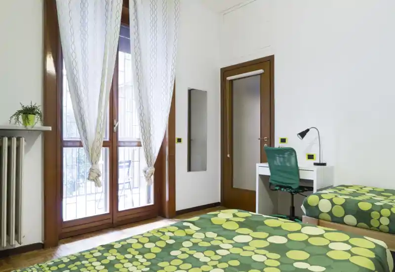 Chambre lumineuse et soignée avec deux lits simples aux housses vertes à motifs, un petit bureau et une chaise, et de grandes portes-fenêtres donnant sur un balcon qui apportent beaucoup de lumière naturelle.