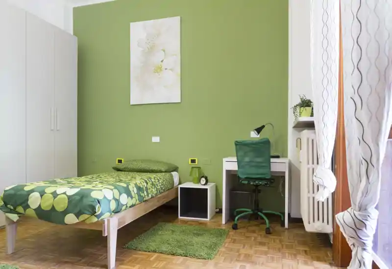 Chambre simple soignée au décor vert, lit en bois, table de chevet et petit bureau près de la fenêtre. Luminosité naturelle et style harmonieux en font un espace accueillant, parfait pour un étudiant ou un invité.